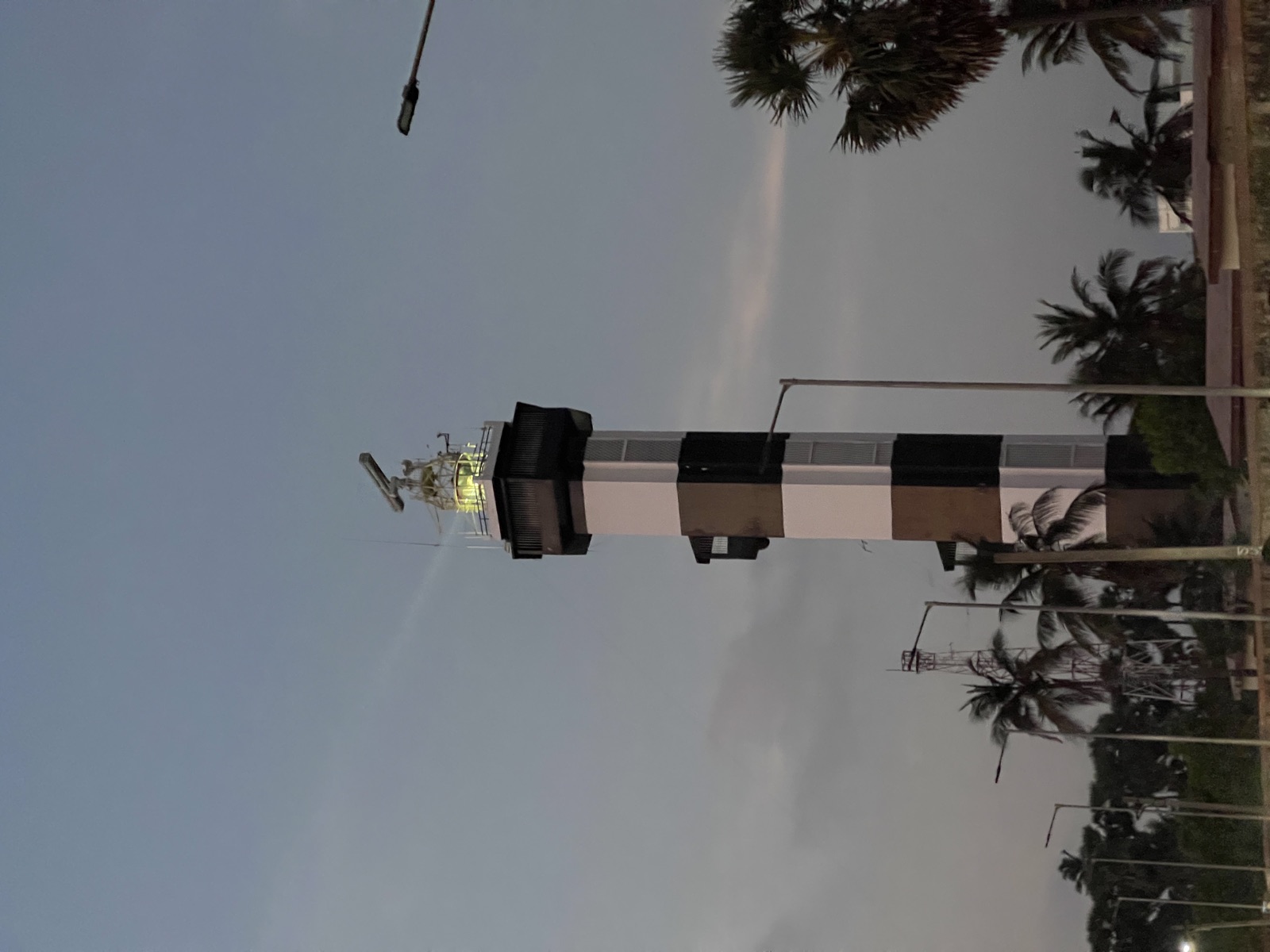 Black and white striped lighthouse at dusk with its beam lit, palm trees below