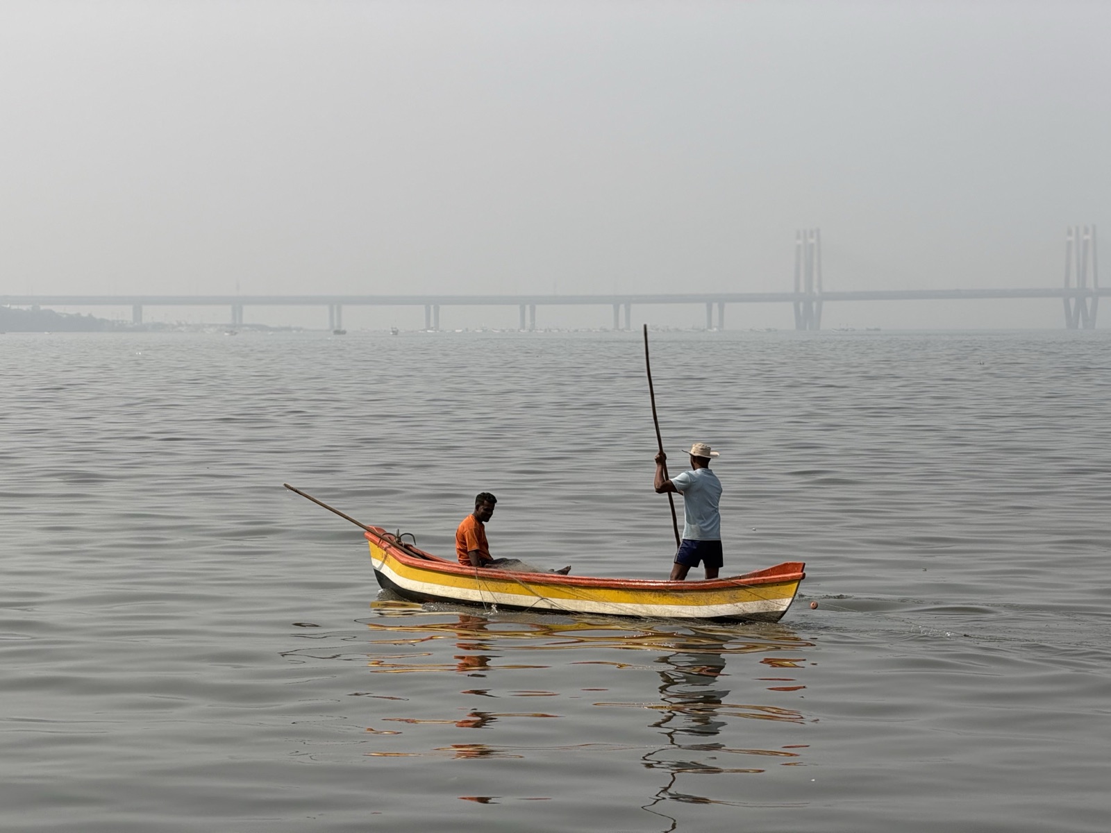 Two fishermen poling a striped wooden boat, Mumbai’s Sea Link bridge hazy behind
