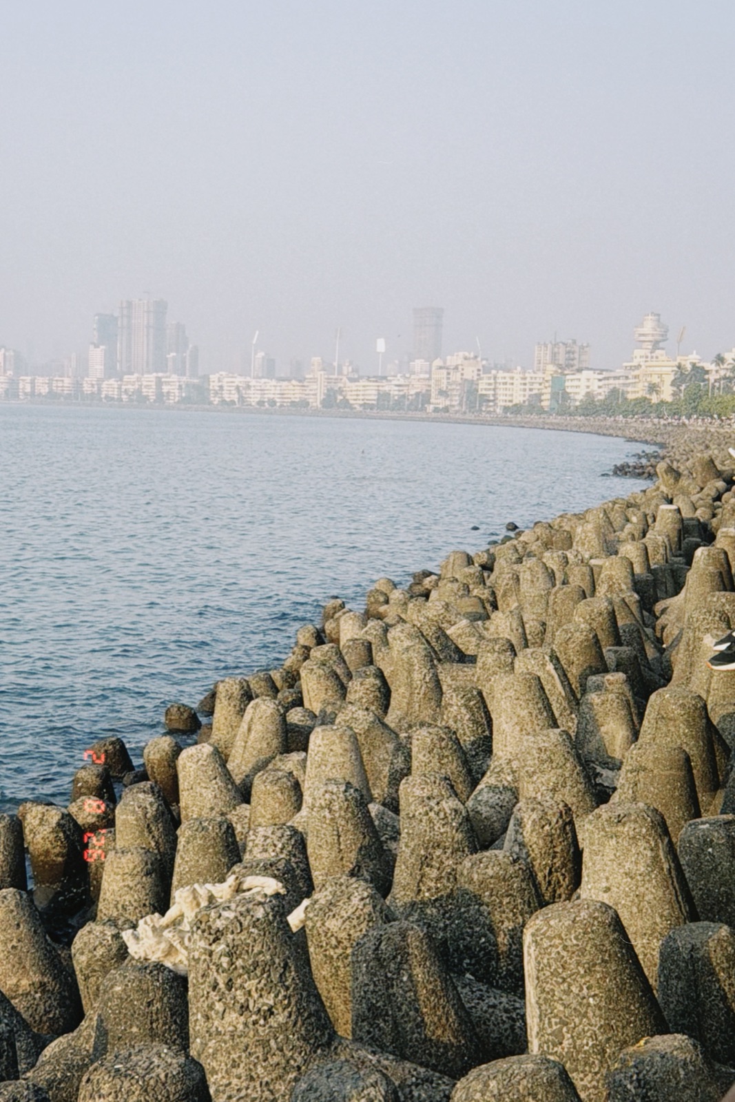 Tetrapod sea wall curving along Marine Drive toward Mumbai’s hazy skyline, film grain look
